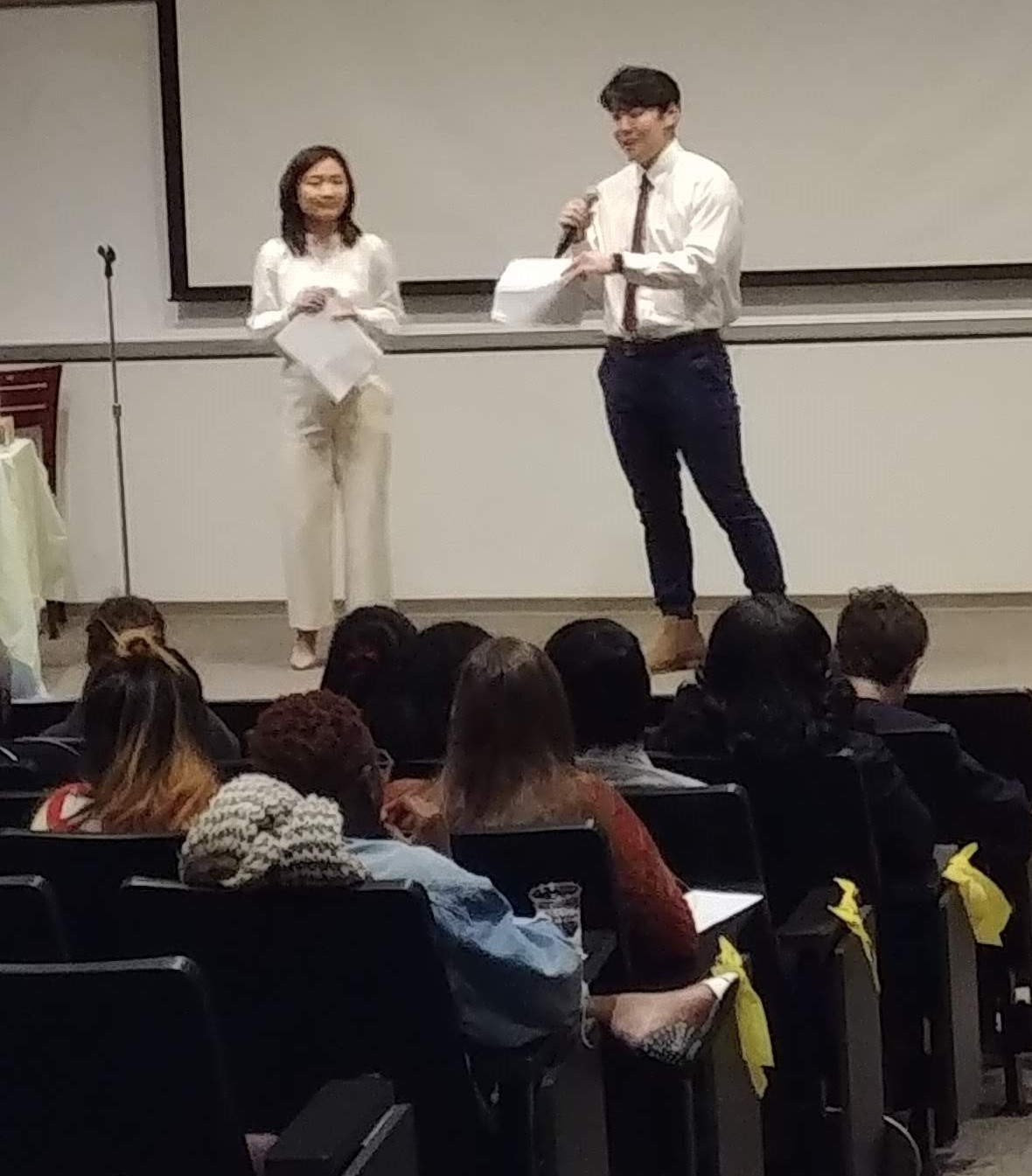 Photo of the 2018 Screening. The two hosts stand at the front of an auditorium in front of a blank screen facing the audience.