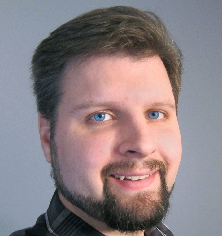 Bio headshot of Brian De Tagyos, a caucasian male with blue eyes, brown hair, beard, and mustache. He stands in front of a blue-grey background wearing a plaid button shirt with a collar, and is smiling directly at the camera.