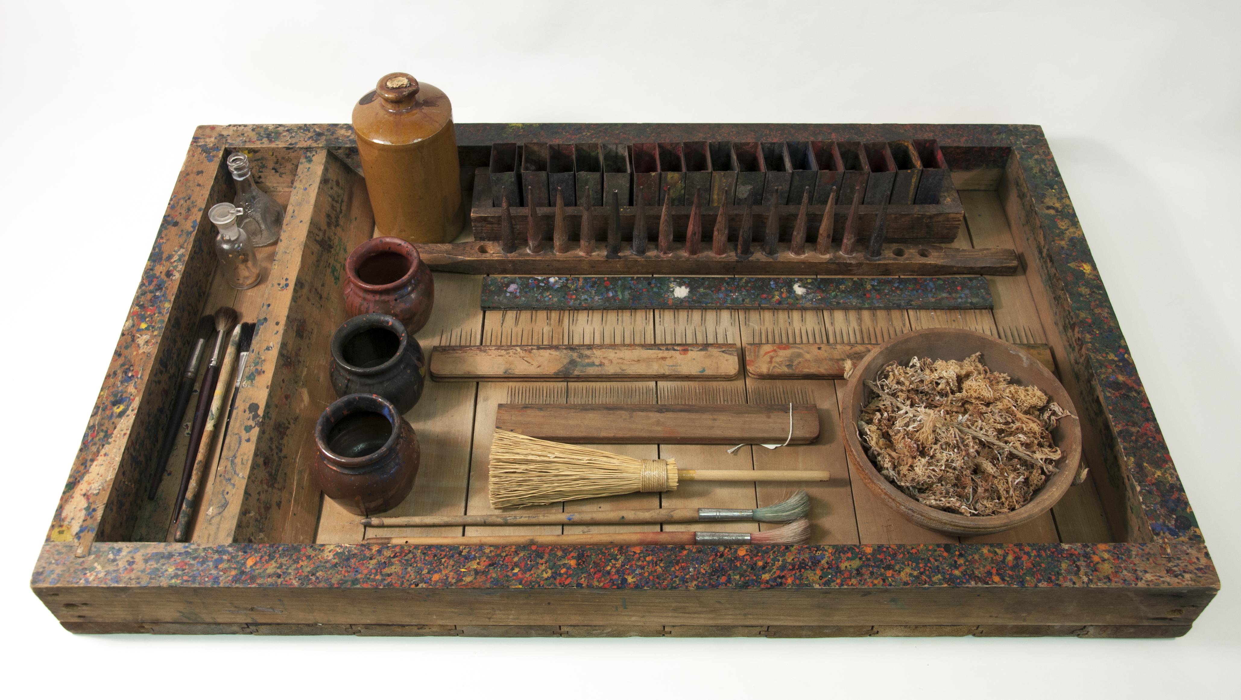 A shallow wooden box containing small jars and other marbling tools, all appearing to be part of a set.